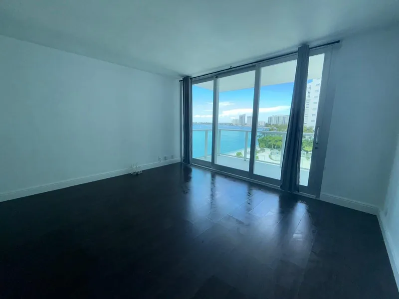 Habitación vacía con suelo de madera oscura y puertas de cristal al balcón con vistas al agua.